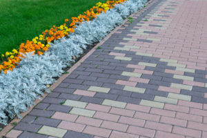 Multicolored driveway design with rectangular shapes of cream, brown, and red. Along paved walkways planted flowerbed with beautiful orange and yellow flowers. In the background, lawn with green grass.